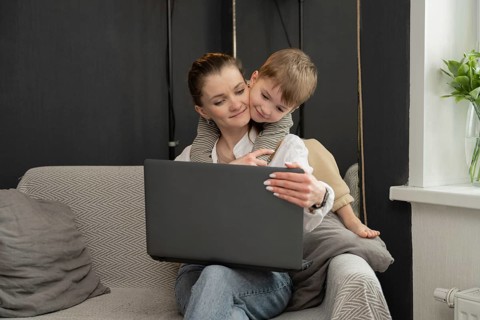 A mother is sitting on a couch using a laptop, while her young child affectionately hugs her from