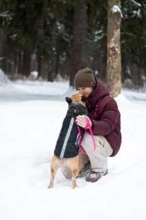 A girl in a warm coat kneels in a snowy forest, sharing a tender moment with their dog. Both enjoy