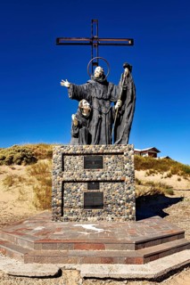Statue of monks with cross in a sandy landscape under clear sky, monument by Francisco De Paola in
