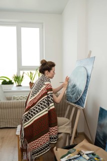 An artist, wrapped in a colorful blanket, paints on a circular canvas in a bright home studio,