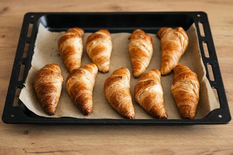 A batch of golden brown, flaky homemade croissants arranged on a parchment lined baking tray. The