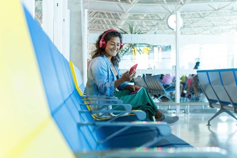 A smiling traveler sits on a colorful airport bench, wearing headphones and holding a smartphone,