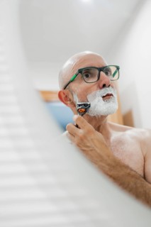 A mature man is seen shaving in a well-lit bathroom, using a razor while looking in the mirror. The