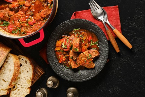 A bowl of beef stew sits on a black surface next to crusty bread. The stew has meat, vegetables,