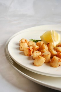The plate displays cooked scallops arranged neatly. A wedge of lemon sits beside the scallops.