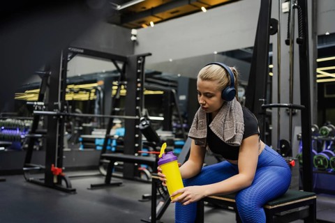 A blonde woman in athletic wear relaxes on a bench in a modern gym, holding a yellow protein shaker