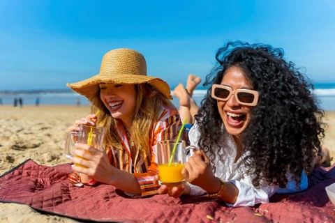 Two cheerful young women lying on a beach blanket, laughing and sipping cold drinks under bright