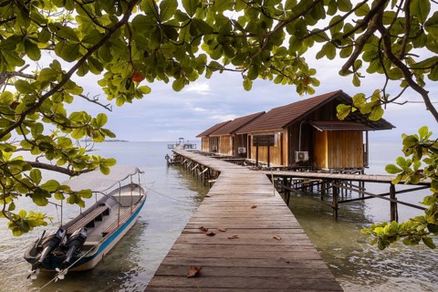 A serene view of wooden huts on stilts and a moored boat framed by lush leaves on Waigeo Island,