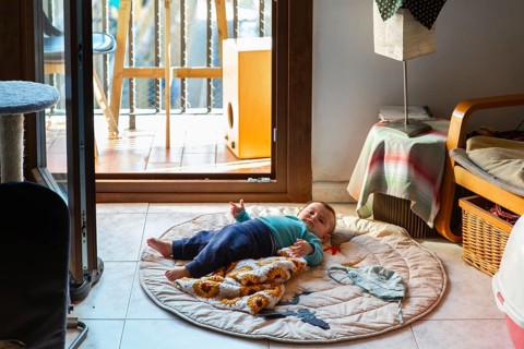 A baby comfortably lies on a round mat in a cozy home near an open balcony door. Natural light
