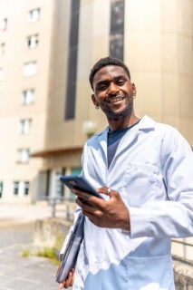 Vertical photo of a cheerful young african doctor using phone standing outside the hospital