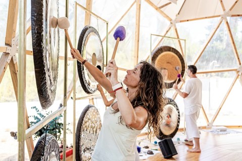 Two sound therapists engage in a gong bath session, striking large gongs with mallets. Their eyes