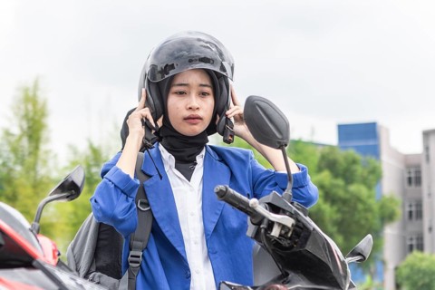 A young Asian Muslim woman wearing a blue jacket and helmet stands by her scooter, getting ready