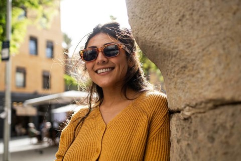 A cheerful Latin woman wearing sunglasses and a mustard sweater smiles warmly, standing against a