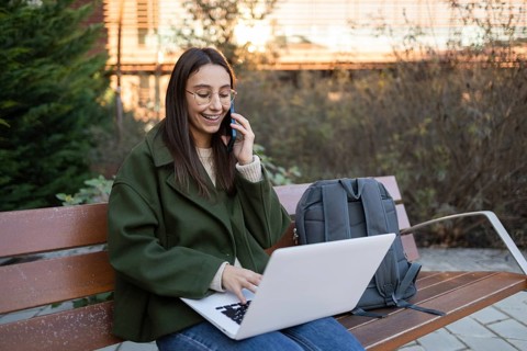 A woman in a green jacket sits on a bench, engaging in conversation on her phone while using a