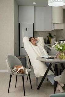 A woman relaxes in a modern kitchen, smiling while sitting at a table. A small dog sleeps on a