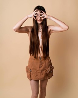 A young woman stands against a beige background in a studio, playfully covering her eyes with her