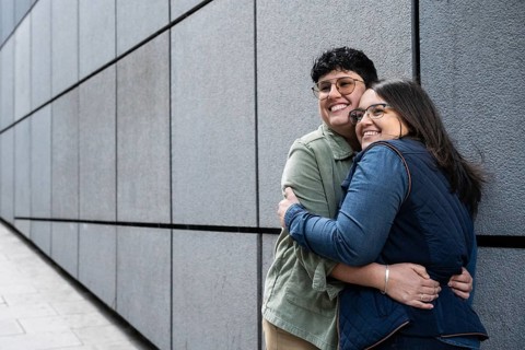 A joyful lesbian couple shares a warm embrace outdoors, standing against a modern urban wall. The