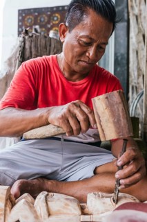 An Indonesian man intricately carves a wooden sculpture using traditional tools. His focus and