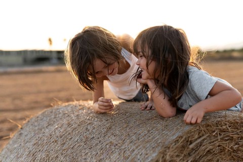 Two smiling siblings enjoy playful moments on a hay bale in a golden countryside sunset. Their