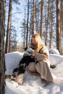 A redhead woman lovingly embraces her Border Collie in a winter forest setting. Sunlight filters