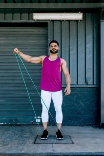 Fit man performs outdoor resistance band workout. Wearing vibrant tank top and leggings, he