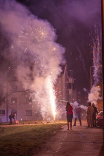 Fireworks light up the night near residential buildings while spectators stand in the street,
