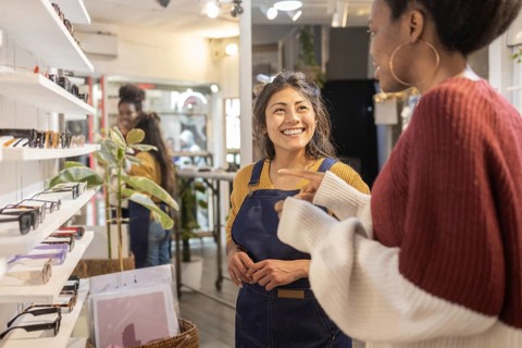 A cheerful sales assistant engages with a customer in a lively sunglasses store. The shelves are