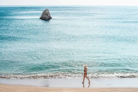 Unrecognizable woman in a summer hat and swimsuit walks leisurely along the shore of beach Gerontas