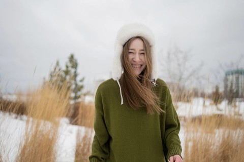A joyful woman in a green sweater and warm white winter hat is smiling in a snowy park. Tall grass