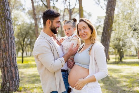 A pregnant woman joyfully stands with her family in a sunny outdoor setting, capturing a moment of