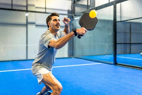 Man in athletic gear striking a pickleball on a blue indoor court, mid action with paddle raised,