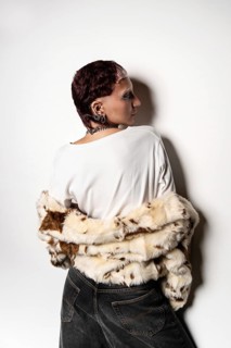 A woman stands confidently with a stylish fur jacket draped over their shoulders, bold earrings,