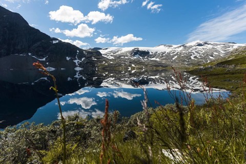 A pristine mountain lake in Norway reflects snow-capped peaks and a bright blue sky Surrounded by