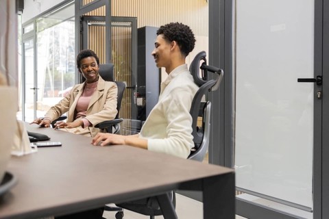 Two colleagues converse in a modern office space, seated comfortably in ergonomic chairs. The