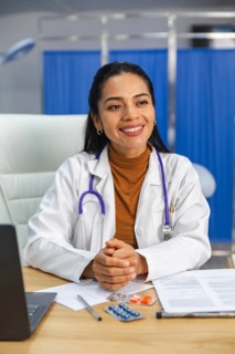 A smiling doctor with a stethoscope around her neck sits at a desk with paperwork and medication on