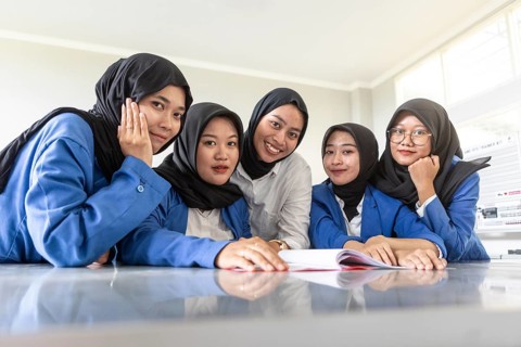 Group of Asian Muslim women sitting together in classroom, wearing hijabs and blue uniforms. They