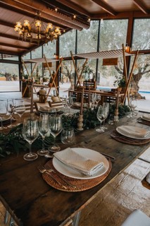 A rustic wooden banquet table with woven placemats, elegant glassware, striped napkins, and lush