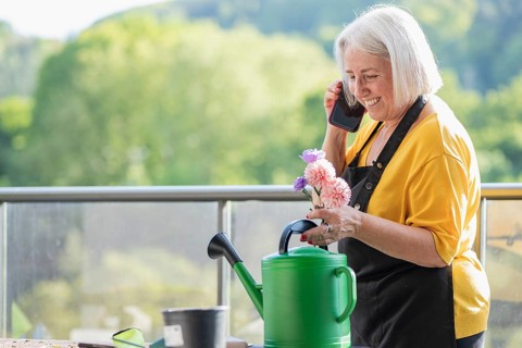 A cheerful elderly woman in a yellow shirt and apron holds colorful flowers while speaking on her