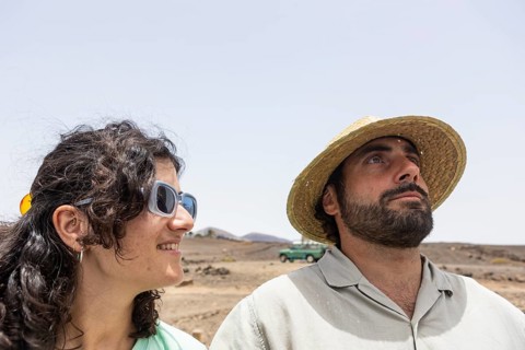 A blind man and woman stand together in a desert terrain. The man is wearing a straw hat and an old