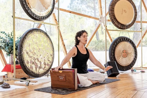 A female sound therapist plays a shruti box with her eyes closed, emphasizing tranquility. Gongs