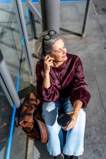 Mature woman with grey hair sits on a city sidewalk, soaking up warm sunlight, holding a cup of