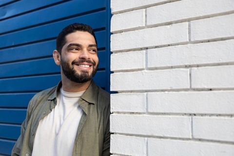 A man with a beard smiles while leaning against a white brick wall of the city, confidently