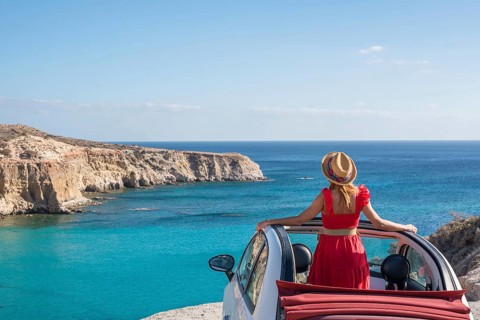 A woman in a red dress stands beside a convertible, overlooking the turquoise waters and rugged