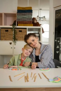Teacher and student sitting at a table, making crafts with colored clay and sticks, creating a warm