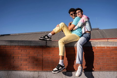 A joyful young gay couple sits on a brick wall, embracing and smiling under a clear blue sky. Their