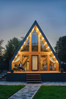 Woman sits on terrace outside a modern triangle log cabin, enjoying the peaceful evening surrounded