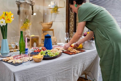 A woman in a green dress arranges a buffet table with an array of vibrant fruits, cured meats, and
