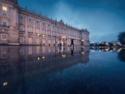 A stunning view of the majestic Royal Palace of Madrid reflecting on wet pavement at dusk. A lone