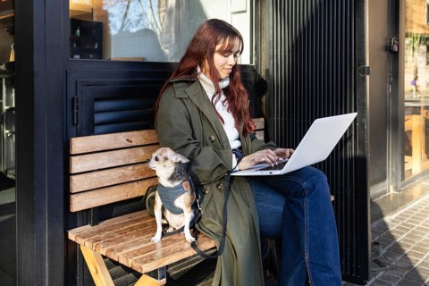 Woman seated on a wooden bench, typing on her laptop outside a cafe. Her small chihuahua sits