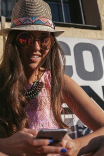 Stylish young woman in a summer hat and sunglasses, smiling while looking at her phone. She sports